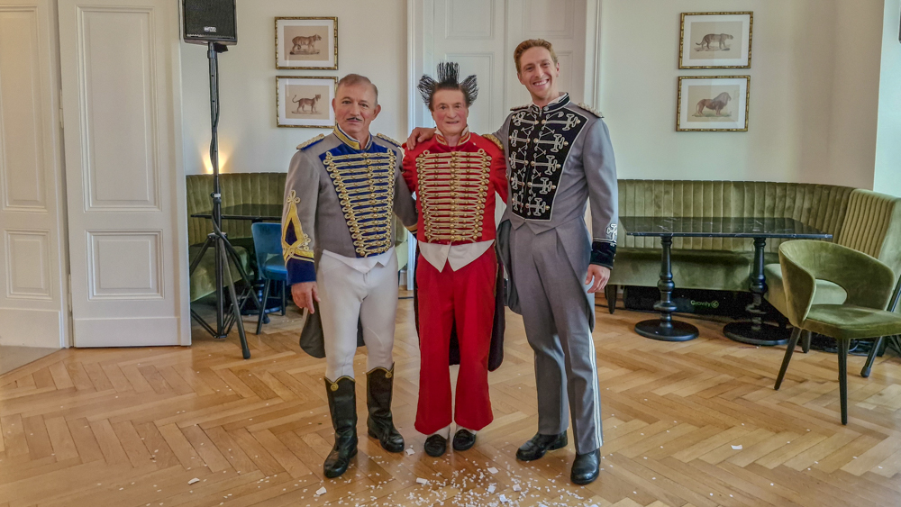 Die Fumagalli Clowns, Gianni Huesca in der Mitte, link sein Bruder Daris und rechts sein Sohn Nick. Die drei stehen in festlichen Uniformen in einem eleganten Raum mit Holzboden. Daris trägt eine graue Uniform mit blauen und goldenen Verzierungen, Gianni eine rote Uniform mit goldenen Akzenten, die Haare hochgegelt, und Nich hat eine graue Uniform mit silbernen Details. Im Hintergrund sind grüne Sitzbänke und an der Wand Bilder von Wildtieren zu sehen. Die Stimmung wirkt freundlich und entspannt.