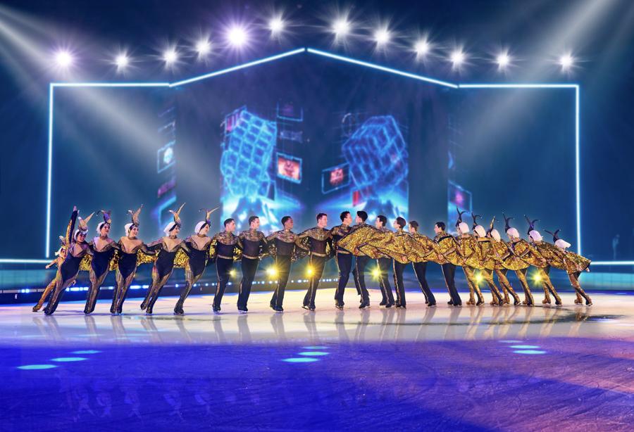 Holiday on Ice Eine große Gruppe von Eiskunstläufern führt eine synchrone Choreografie auf einer beleuchteten Eisfläche auf. Die Läufer sind in glänzende, gold-schwarze Kostüme gekleidet und bilden eine lange, zusammenhängende Linie. Im Hintergrund ist eine futuristische, blau erleuchtete Kulisse mit geometrischen Formen und Lichteffekten zu sehen. Die Szene wird von mehreren hellen Scheinwerfern beleuchtet, die die Performance in den Fokus rücken.