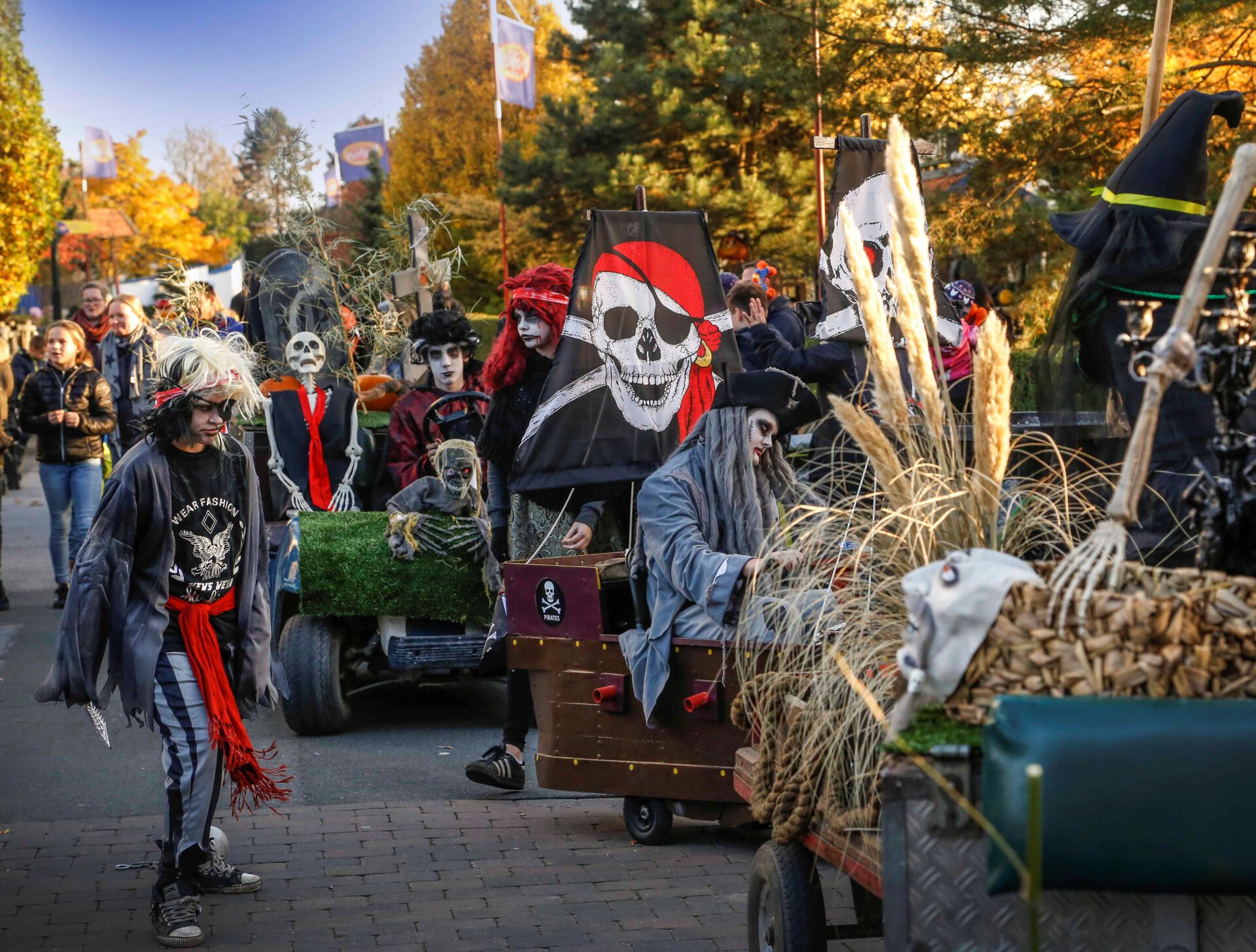 Eine Gruppe von Menschen in Halloween-Kostümen nimmt an einem Umzug im Freien teil. Die Teilnehmer sind als gruselige Figuren verkleidet, darunter Piraten und Skelette, und einige tragen geschminkte Gesichter. Im Vordergrund stehen zwei Personen, von denen ein rot-schwarzer Schal und gestreifte Hosen trägt. Im Hintergrund sind Wagen mit Dekorationen wie Totenkopffahnen und ausgestopften Figuren zu sehen. Die Umgebung zeigt herbstliche Bäume und einige Zuschauer, die den Umzug beobachten. Die Szene wirkt festlich und thematisch passend zu Halloween.