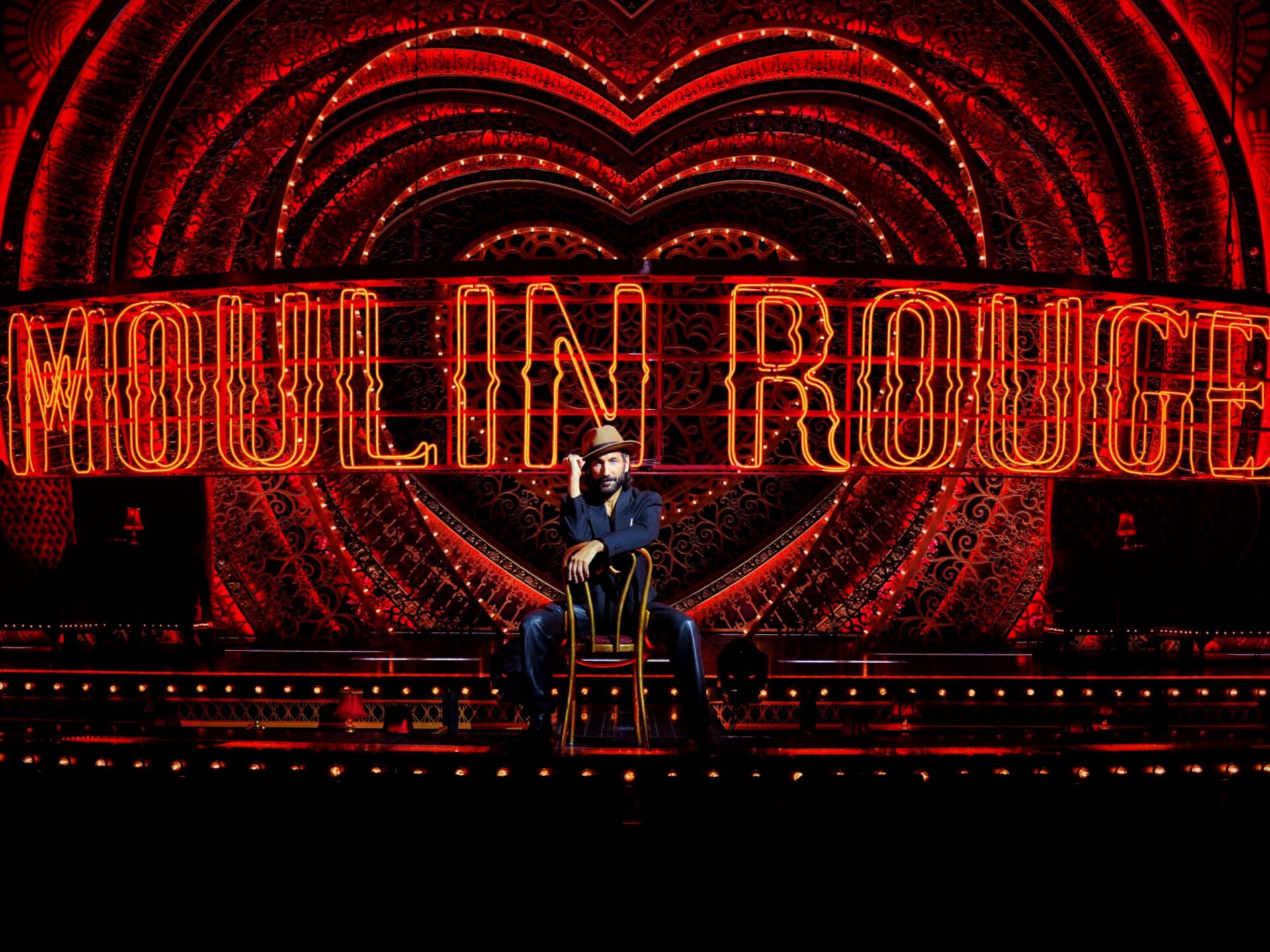 Moulin Rouge! Das Musical, Massimo Sinato sitzt in einem eleganten Anzug auf einem Stuhl vor einem spektakulär beleuchteten Hintergrund, der in leuchtendem Rot die Worte "Moulin Rouge" in großen, neonartigen Buchstaben zeigt. Der Hintergrund besteht aus kunstvollen, kreisförmigen Mustern, die sich in konzentrischen Kreisen hinter den Schriftzug einfügen. Der Mann trägt einen Hut und lehnt lässig auf dem Stuhl, während er leicht seinen Kopf berührt. Die Bühne ist in warmem, glühendem Licht getaucht, was die dramatische und glamouröse Atmosphäre verstärkt.