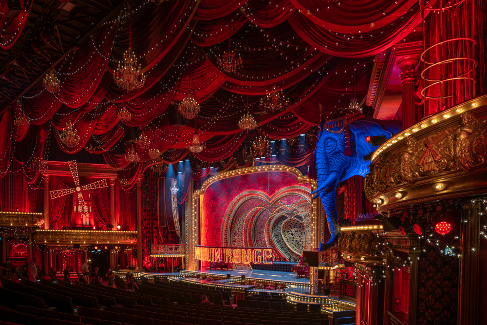 Innenansicht des Moulin Rouge mit einer prächtig dekorierten Bühne, die in rotem Licht erstrahlt. Im Zentrum der Bühne befindet sich ein großes, herzförmiges Bühnenbild aus kunstvollen Ornamenten, umrahmt von einem leuchtenden Bogen. Links ist ein beleuchtetes Windrad zu sehen, und rechts ragt eine große blaue Elefantenstatue hervor. Der Raum ist mit roten Samtvorhängen, funkelnden Kronleuchtern und Lichterketten geschmückt, die eine glamouröse Atmosphäre schaffen. Im Vordergrund sind einige Menschen auf den Sitzreihen zu erkennen.