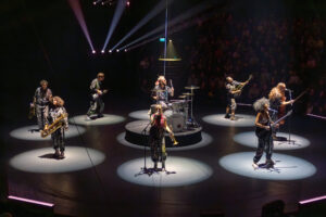 Die siebenköpfige Liveband der Flic Flac X-MAS Show spielt auf einer zentralen Bühne unter Spotlights – mit Saxophon, Schlagzeug, E-Gitarre und Gesang.
