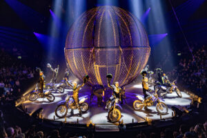 Zehn Motorradfahrer posieren vor dem Globe of Speed in der Manege der Flic Flac X-MAS Show in Dortmund – spektakuläre Stuntshow mit tobendem Publikum.