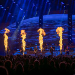 Ein Konzertauftritt mit einer Band auf einer großen Bühne. Im Vordergrund sieht man das Publikum im Dunkeln, während mehrere Flammenwerfer in einer Linie vor der Band aufsteigen. Die Bühne ist mit blauen Lichtern beleuchtet, und im Hintergrund wird ein großes Bild von Wellen auf einem Bildschirm projiziert. Die Bandmitglieder spielen Gitarre und singen, während die Flammen die Szenerie dramatisch erhellen.